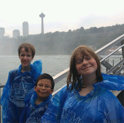A group of three children smile and stand at the railing near Niagara Falls, wearing blue rain ponchos.