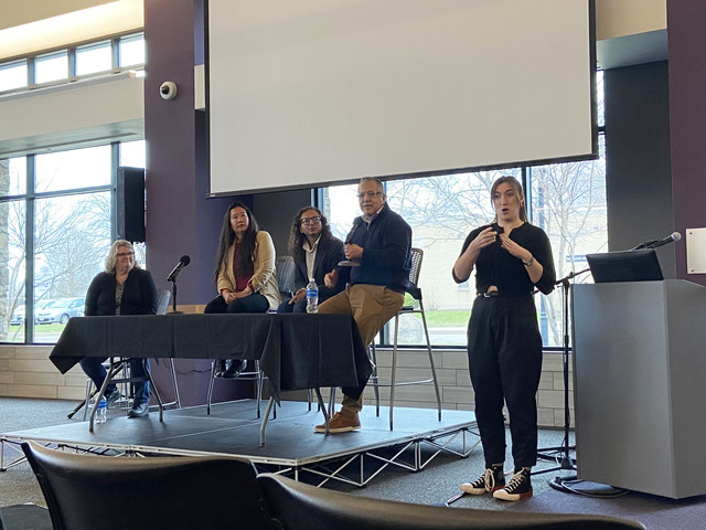 Panel members sit on a small platform stage in front of a retractable film screen. An ASL interpreter stands to the right, relaying their words.
