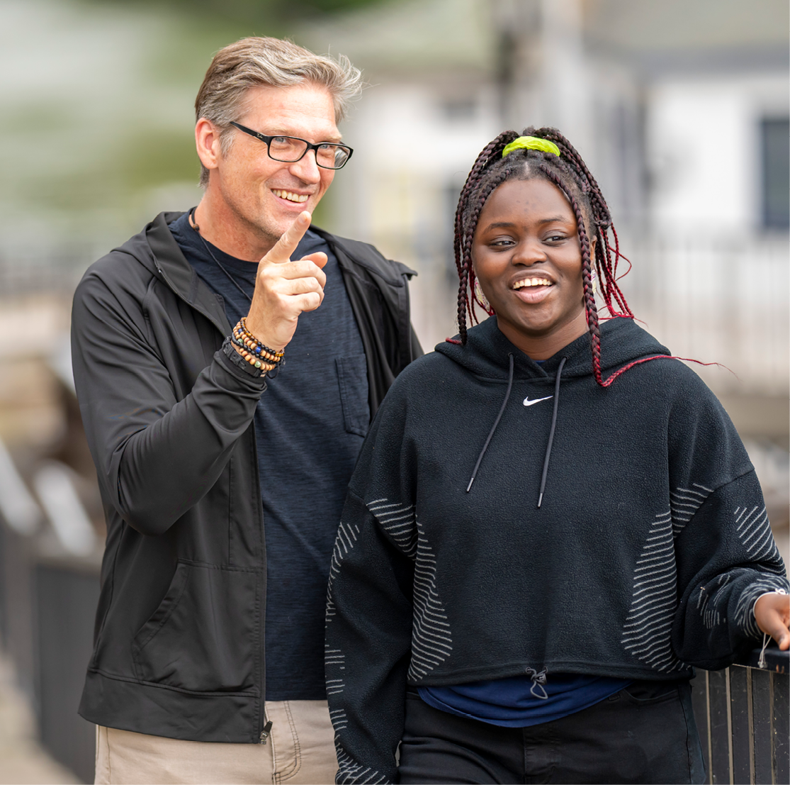 A white man with short blond hair and glasses smiles and points forward, gesturing to a point of interest for the woman he is supporting who stands beside him: a young black woman whose black and red braided hair is pulled into a ponytail. She, too, is smiling. Both wear black jackets.