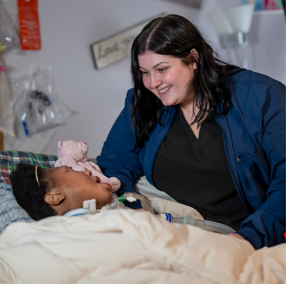 In a home setting, a female nurse with black hair smiles at a patient who is lying in bed, facing her.
