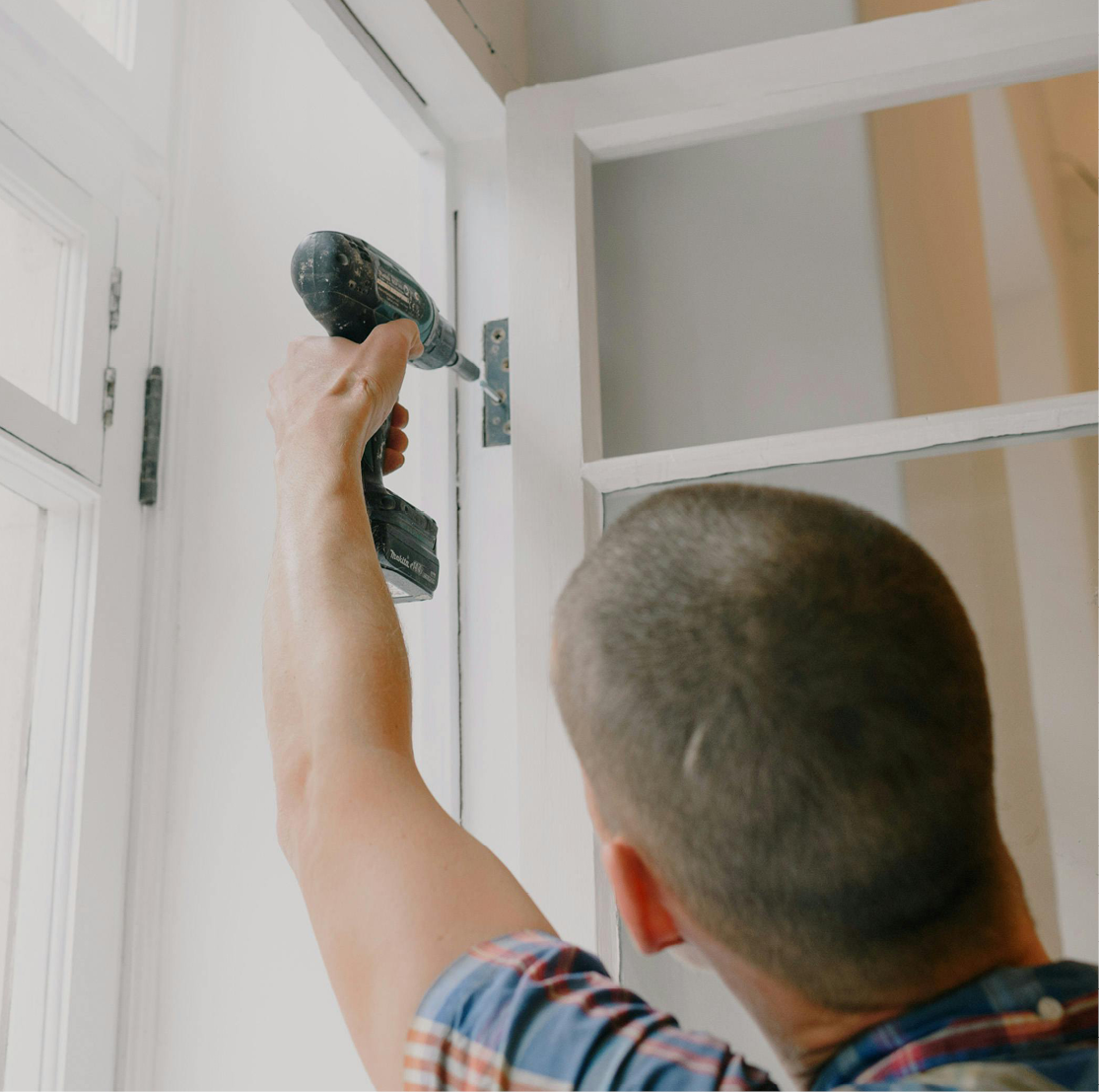 A white man with short shaved hair uses an electric screwdriver to repair a hinge on a glass paned door.
