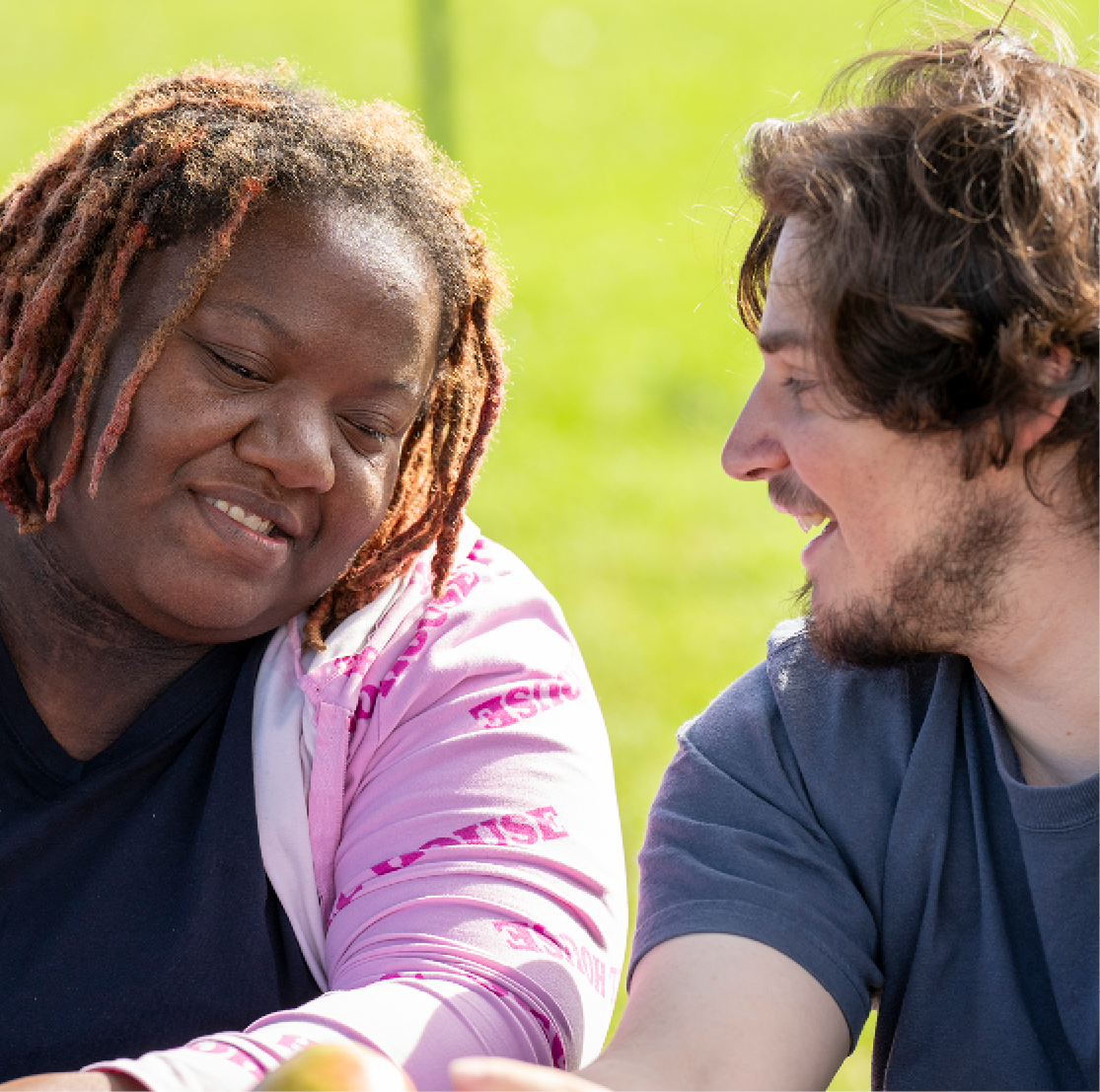 A young woman with short braided hair smiles and speaks with a male employee at an outdoor picnic table.
