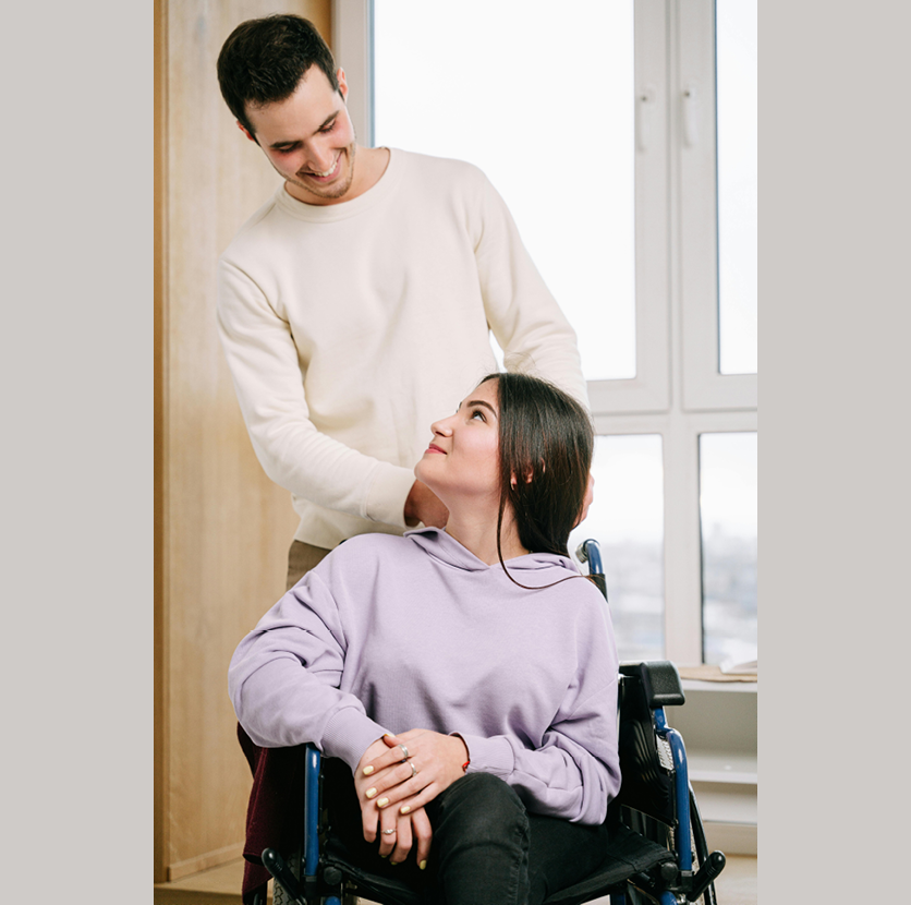 A young man stands in a cream colored room, smiling at a young woman, who returns the smile and sits in front of him in a wheelchair.