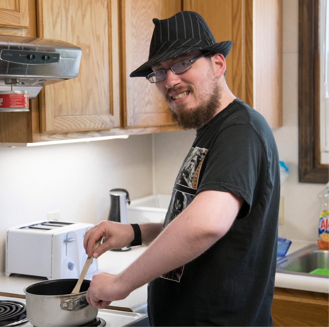 Young man wearing fedora and glasses smiling and cooking from a stovetop pot in a bright apartment kitchen.