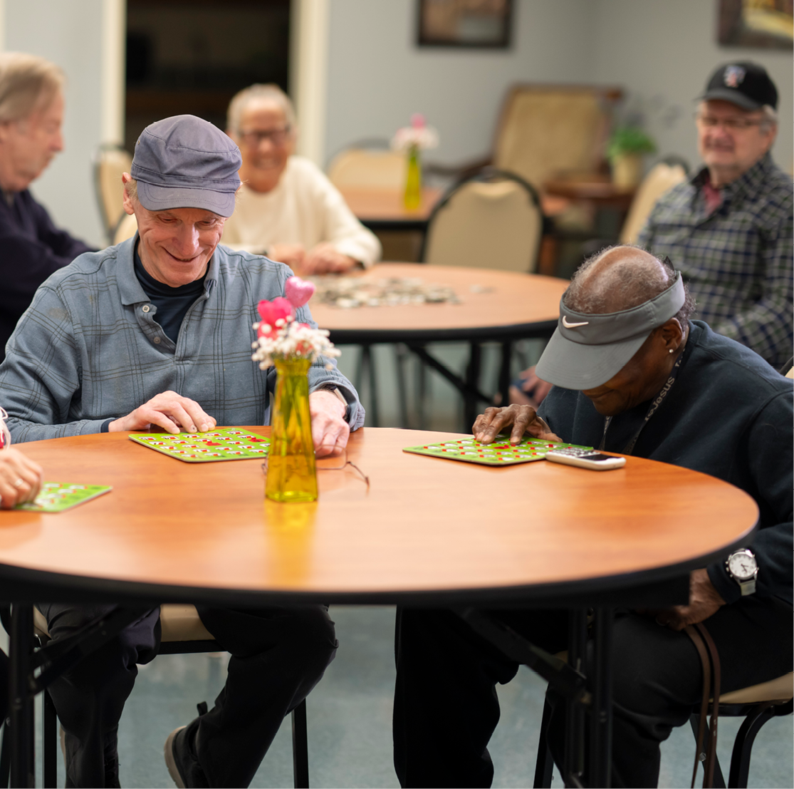 Pair of senior adults smiling and playing bingo at a round community room table.