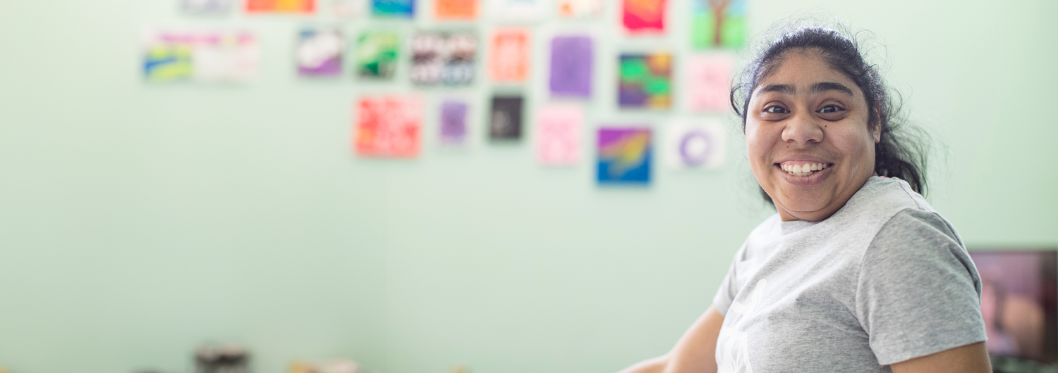 Dark haired young woman smiling with an impression of colorful artwork hanging in the background.