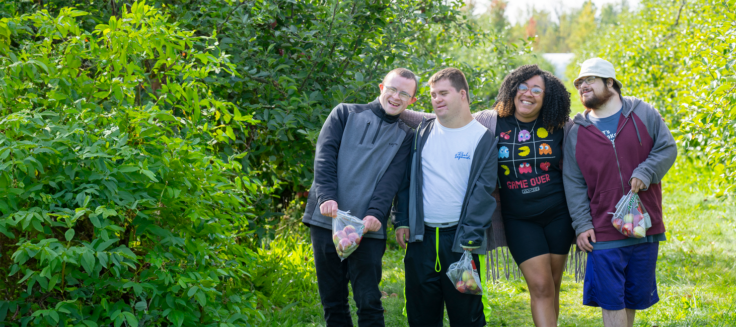 A smiling DSP and three young men she supports walk side-by-side, smiling, through an apple orchard.