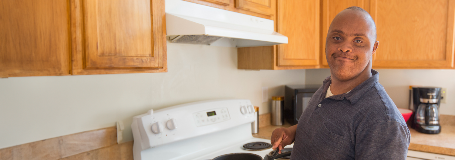 A young Black man with Down syndrome works on cooking skills as he heats a pan on a stovetop.