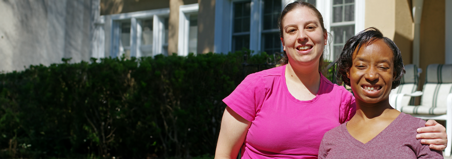 Two women stand together smiling outside of their home on a sunny summer day.