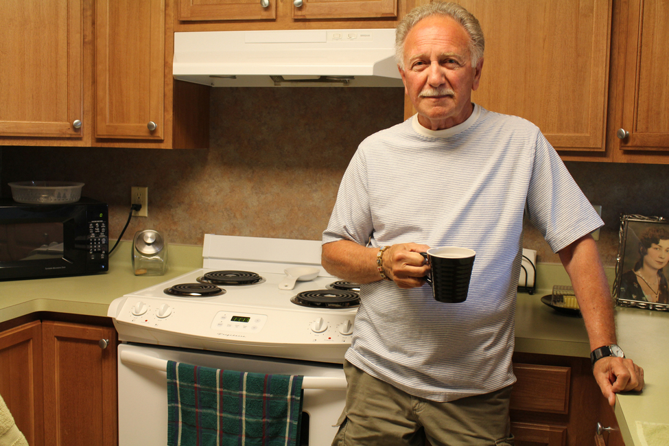 Older man stands in kitchen in front of his stove holding a coffee mug