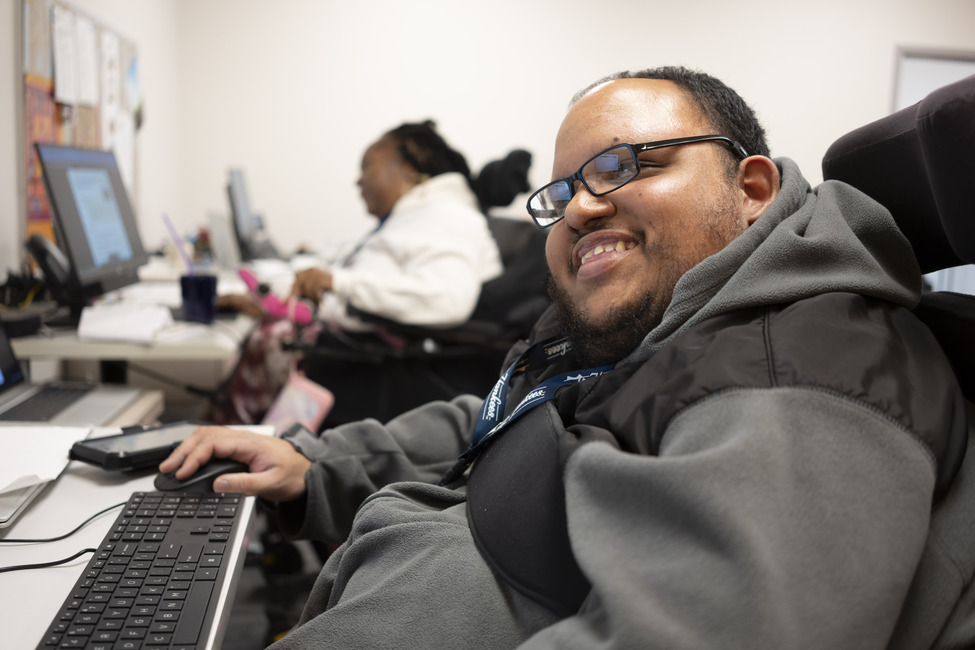 A young Hispanic man in a wheelchair with dark hair and glasses, smiles while using a desktop computer.