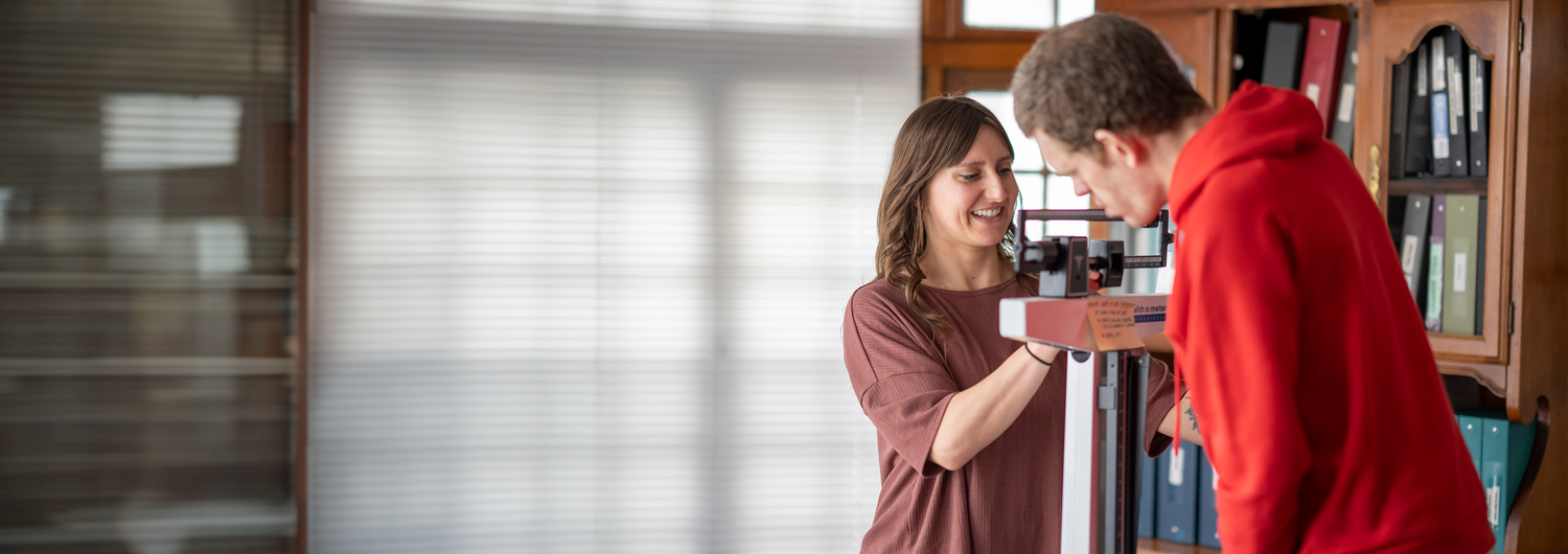 In an in-home office space, a smiling female nurse with shoulder length brown hair assists a young man with sandy brown hair on a doctor's office style scale. 