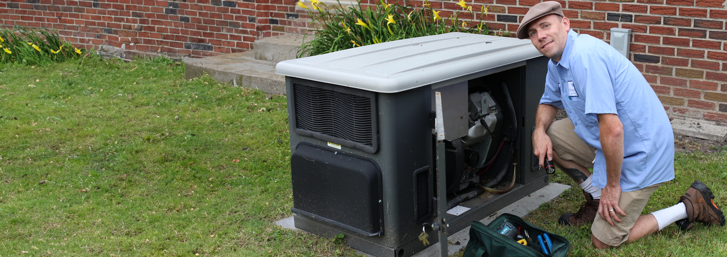 Outside of a group home, a facilities technician stoops beside an electrical unit with a bag of repair tools. He wears a light blue button-down uniform shirt.