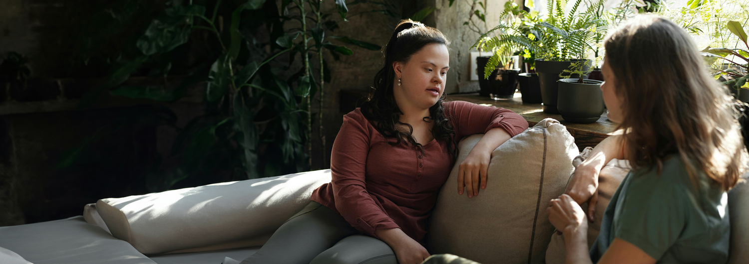 A young woman with Down Syndrome sits on a couch in a plant-filled living room, chatting with another woman who sits beside her on the couch.