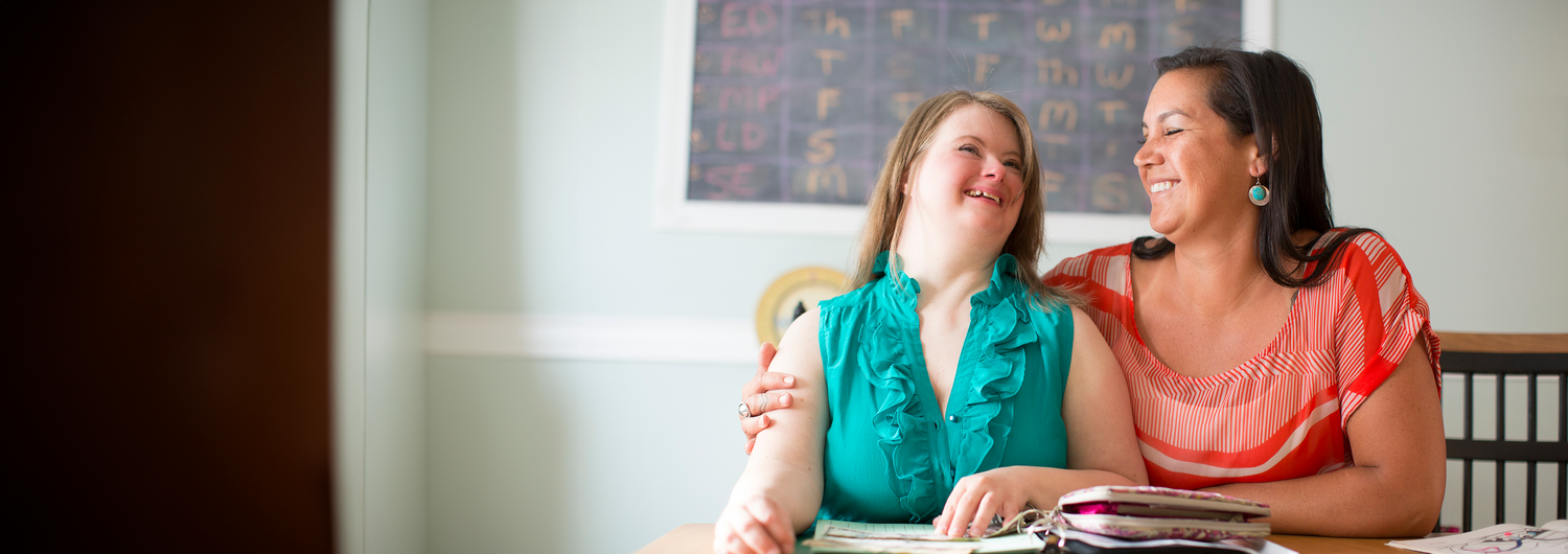 A young woman with Down syndrome smiles and sits at a table. Beside her sits another woman with dark hair, who smiles and embraces her with one arm.