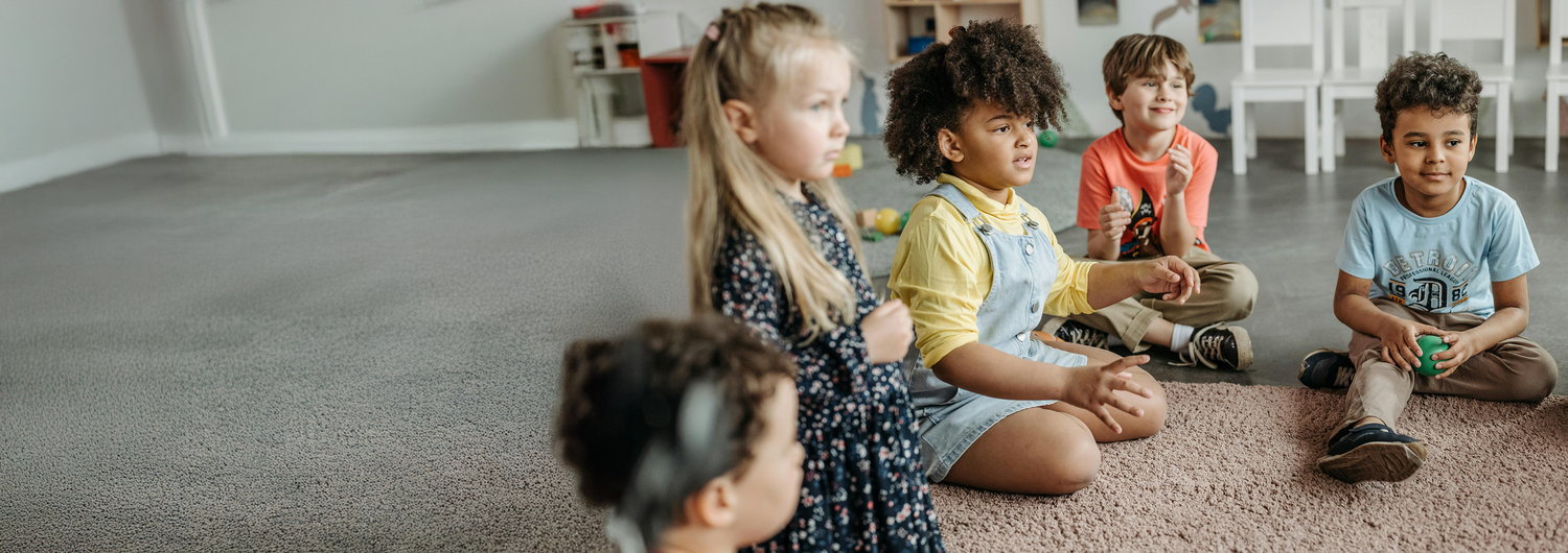 Diverse children sitting together on the floor in a classroom style setting, looking towards an instructor off-screen.
