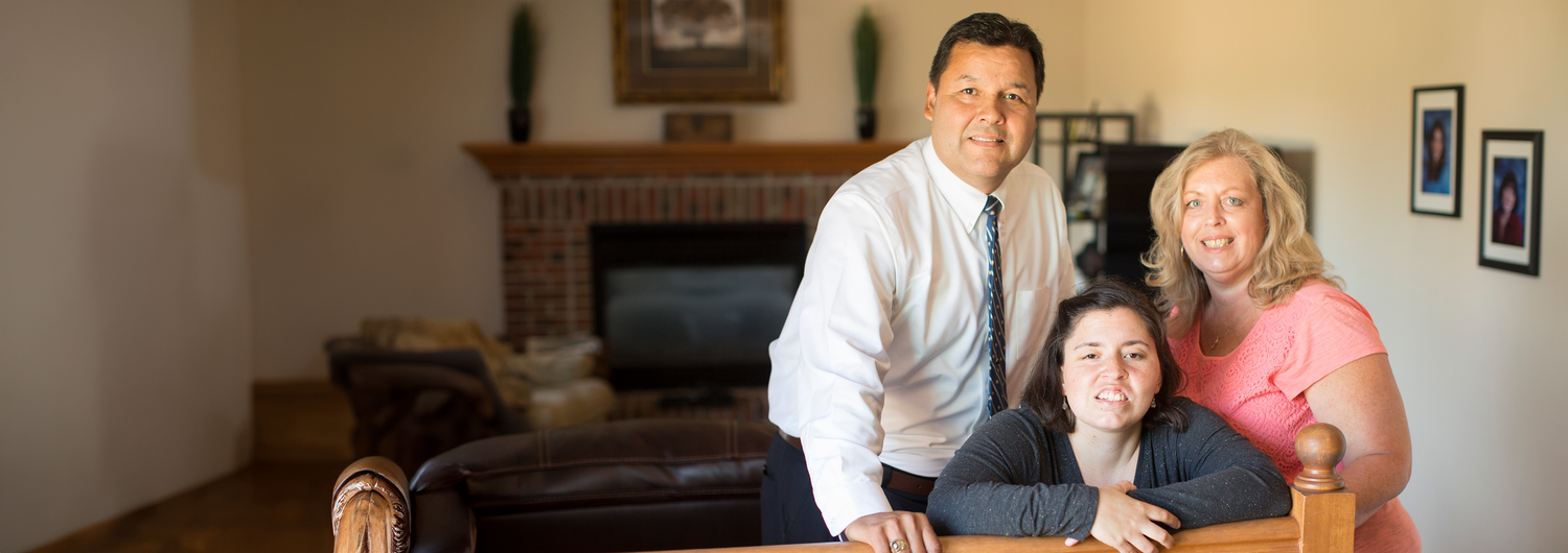 A Hispanic family, a man, woman and their adult daughter smile and pose in a warm living room with cream colored walls and a brick hearth visible.
